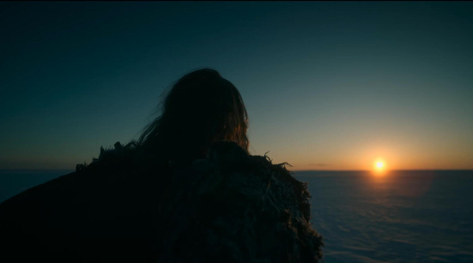 A screencap of the creature from the 2025 Frankenstein film looking towards the sunrise. He is faced away from the viewer.