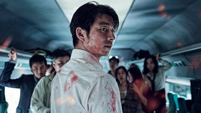 A screencap of Seok-Woo turning slightly towards the viewer with a frown on his face. There are blood stains on his head and shirt. The background includes the interior of a train and other passengers.