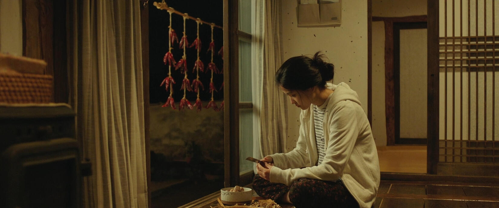 A screencap of Hye-Won sitting on her floor while eating. Chili peppers hang on strings outside her door.